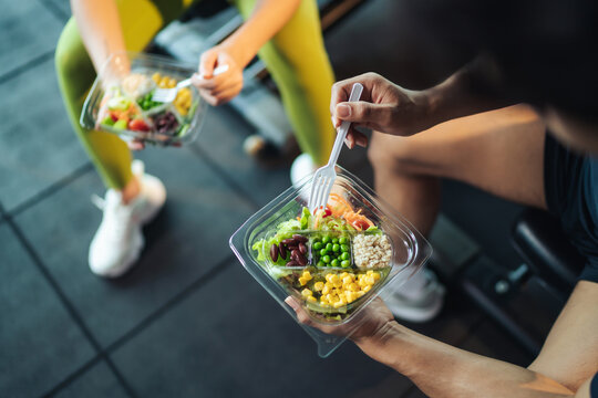 Two lifters eating healthy meal-prep bowls between machines at the gym